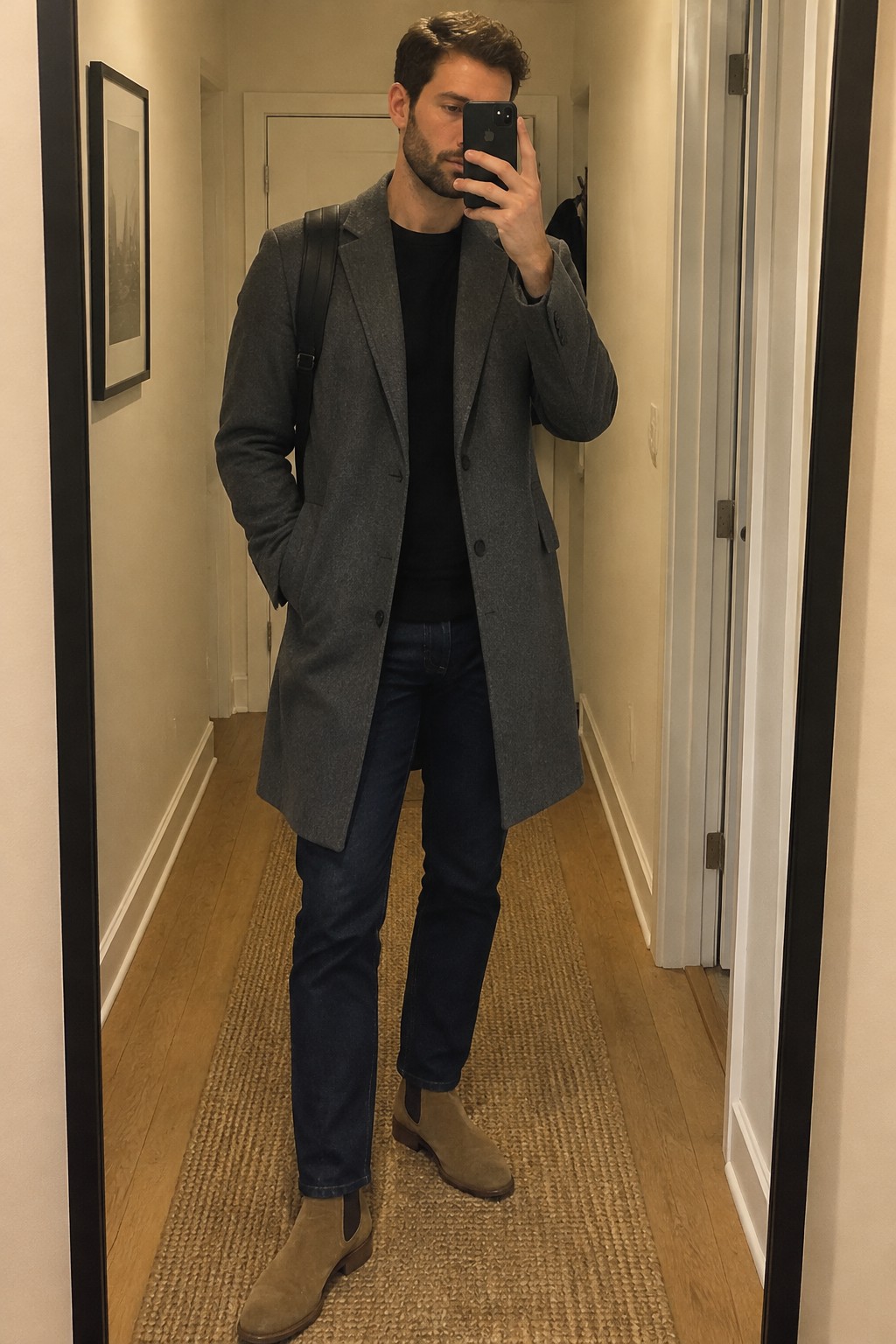 Man wearing a charcoal long wool overcoat over a black crewneck, dark jeans, and tan suede Chelsea boots.