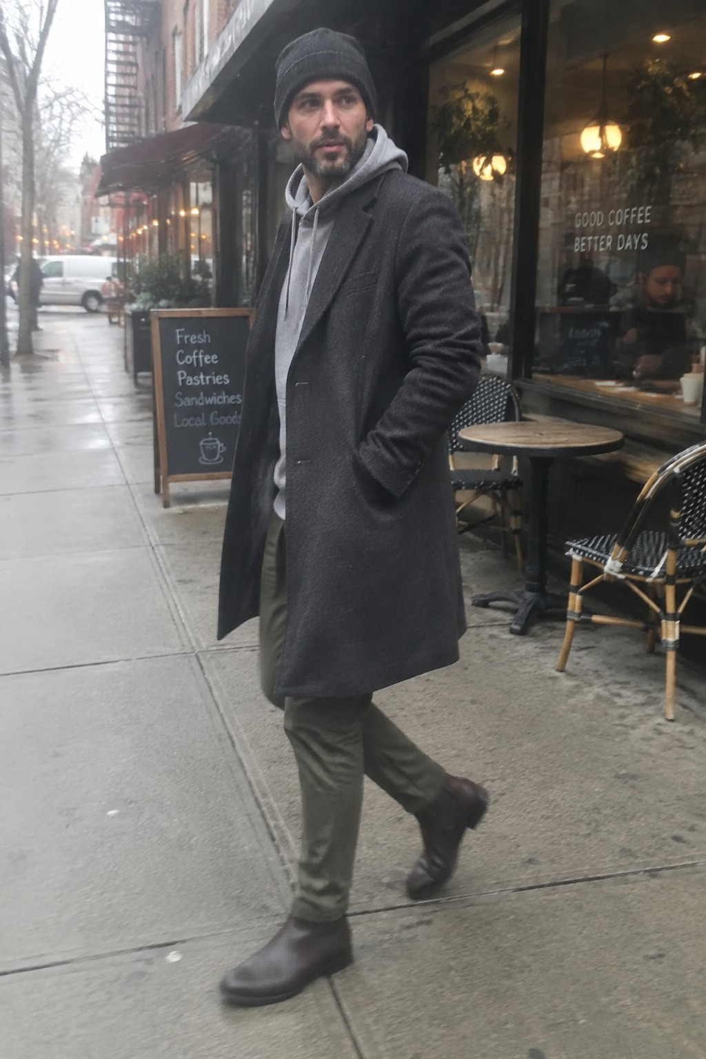 A man wears a charcoal long wool overcoat over a gray hoodie, olive trousers, a dark knit beanie, and dark leather boots.