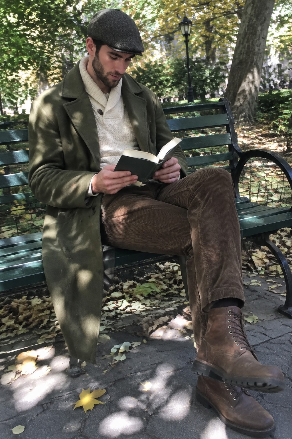A man in an olive long overcoat, cream knit sweater, brown corduroy trousers, dark flat cap, and brown lace-up boots sits on a bench reading a book.