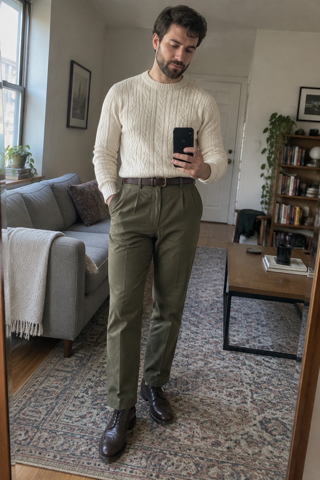 A man takes a mirror selfie wearing a cream cable-knit sweater tucked into olive pleated trousers with a dark brown belt and brown leather lace-up shoes.
