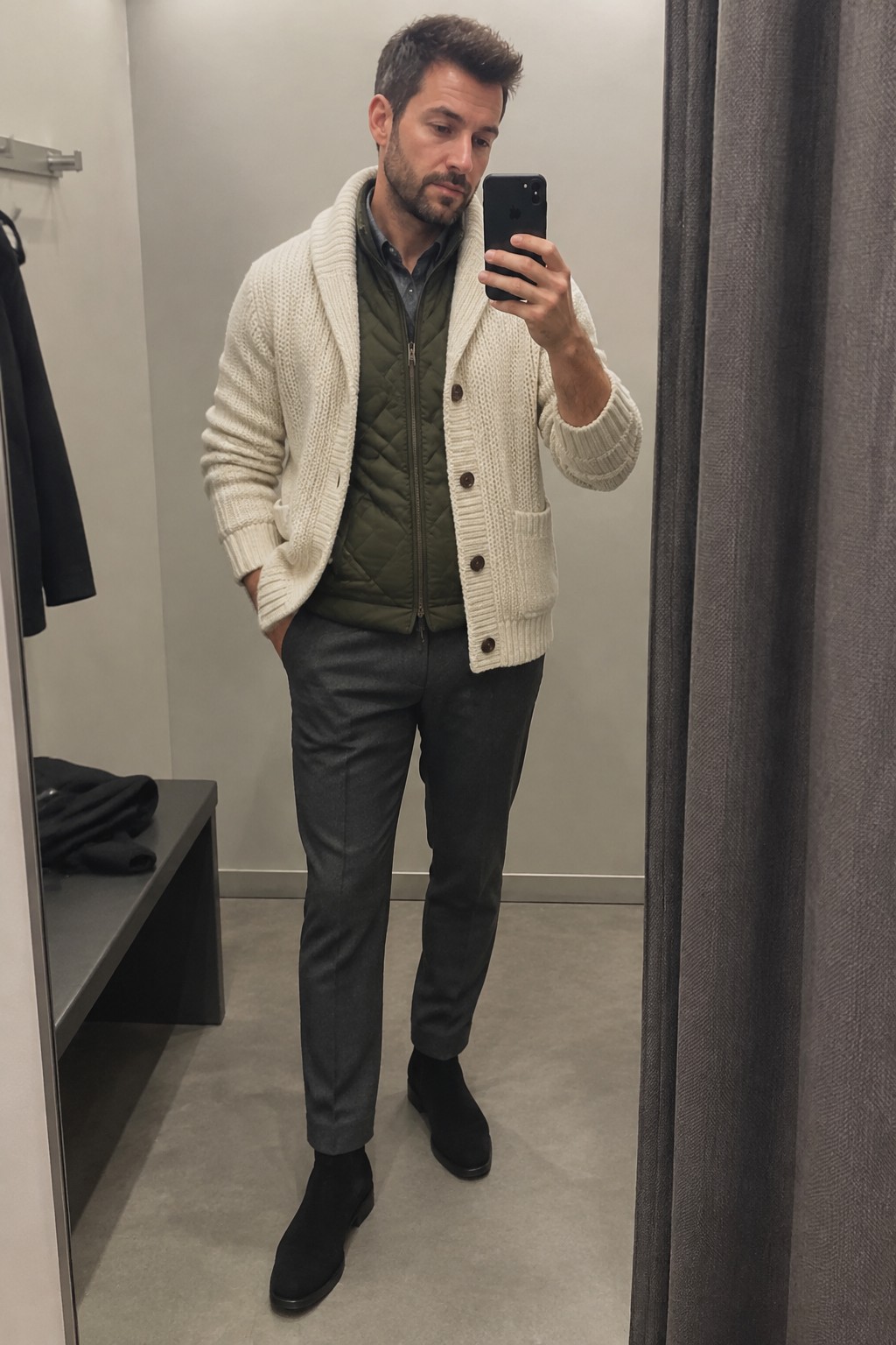 A man wears a cream cable-knit button cardigan over an olive quilted vest and charcoal shirt, paired with dark gray trousers and black ankle boots.