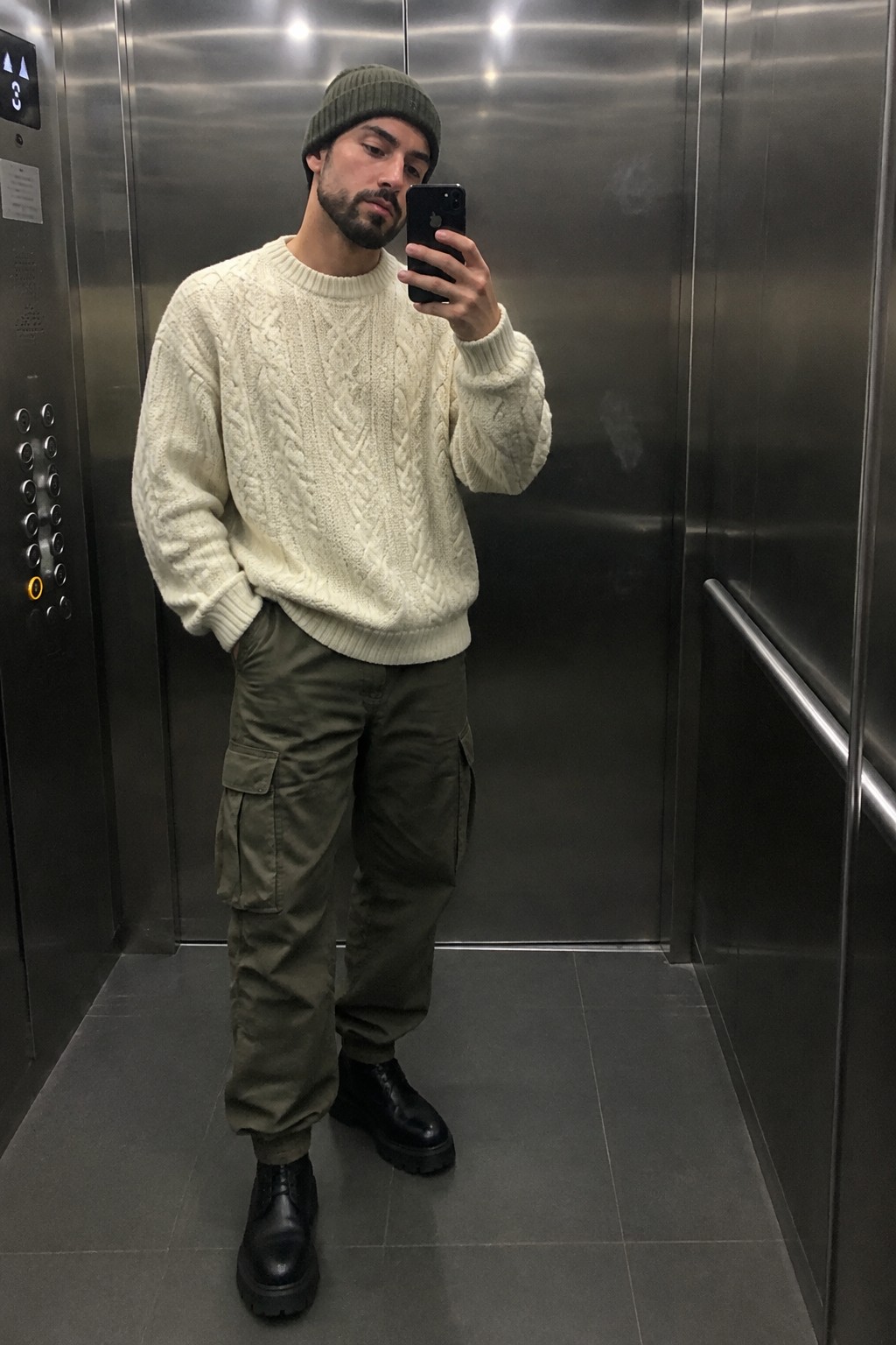 A man wearing a cream cable-knit sweater, olive cargo pants, a dark green beanie, and black lace-up boots.