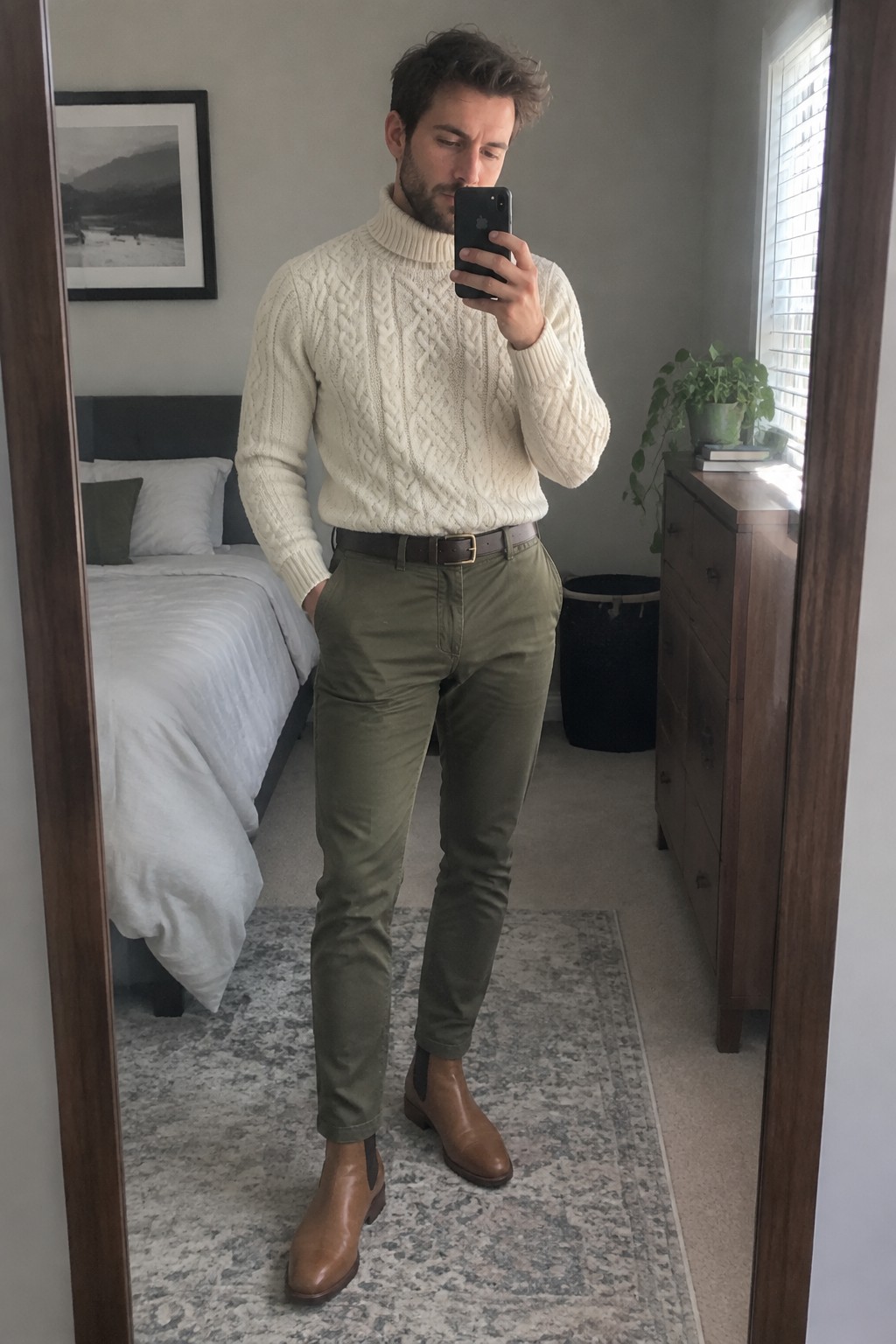 A man wearing a cream cable-knit turtleneck tucked into olive chinos with a brown leather belt and matching Chelsea boots.