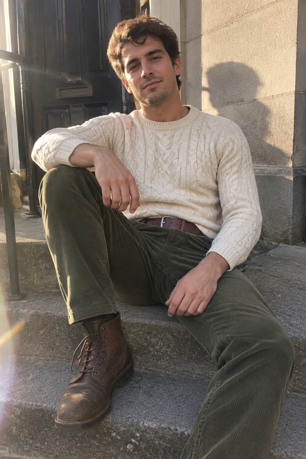 A man wearing a cream cable-knit sweater, olive corduroy trousers, a brown belt, and brown lace-up boots.