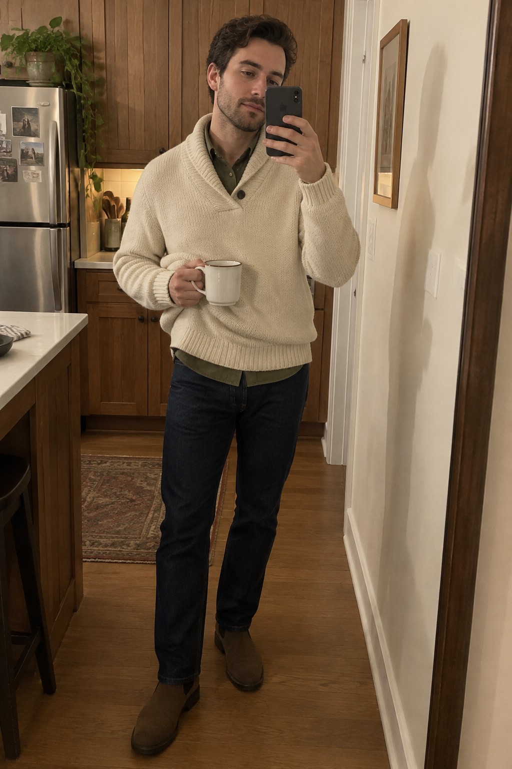 A man taking a mirror selfie in a cream shawl-collar sweater layered over an olive shirt, dark jeans, and brown suede Chelsea boots.