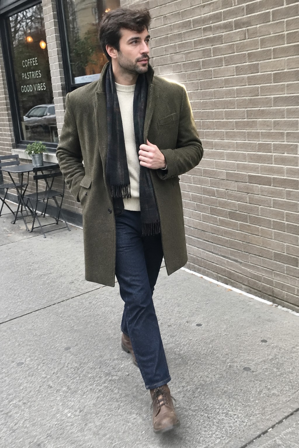A man wearing an olive green long coat over a cream ribbed sweater, a dark scarf, dark blue jeans, and brown lace-up boots.