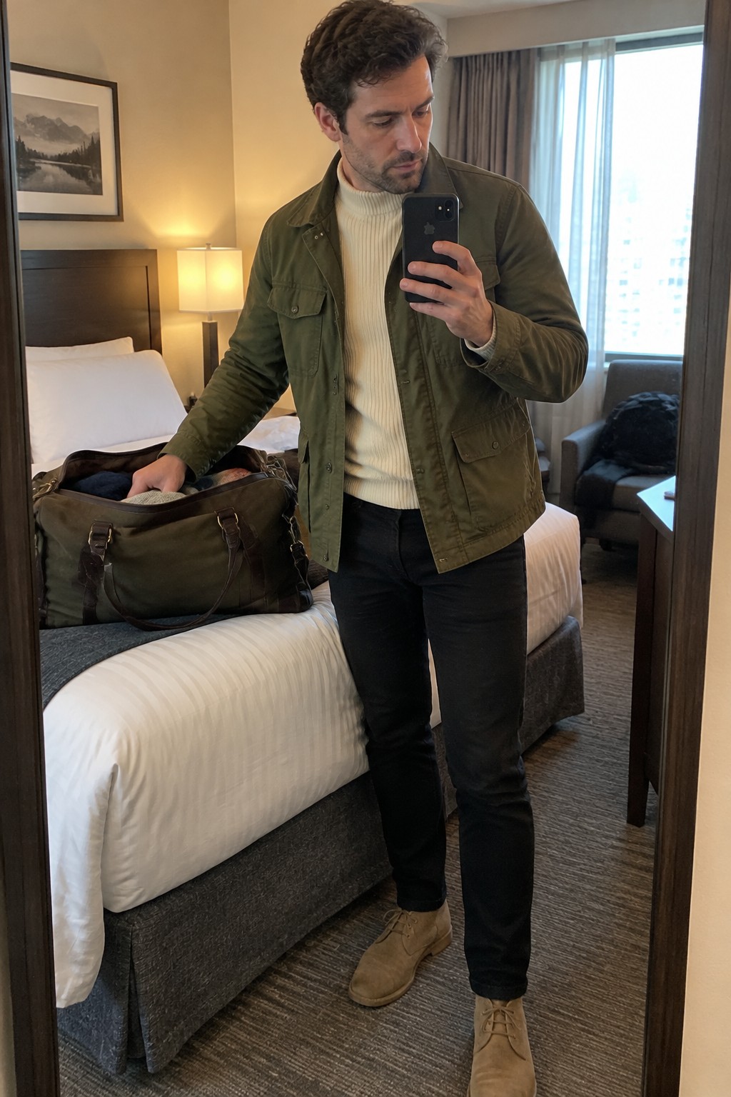Man wearing an olive field jacket over a cream ribbed turtleneck, dark jeans, and tan suede lace-up boots.