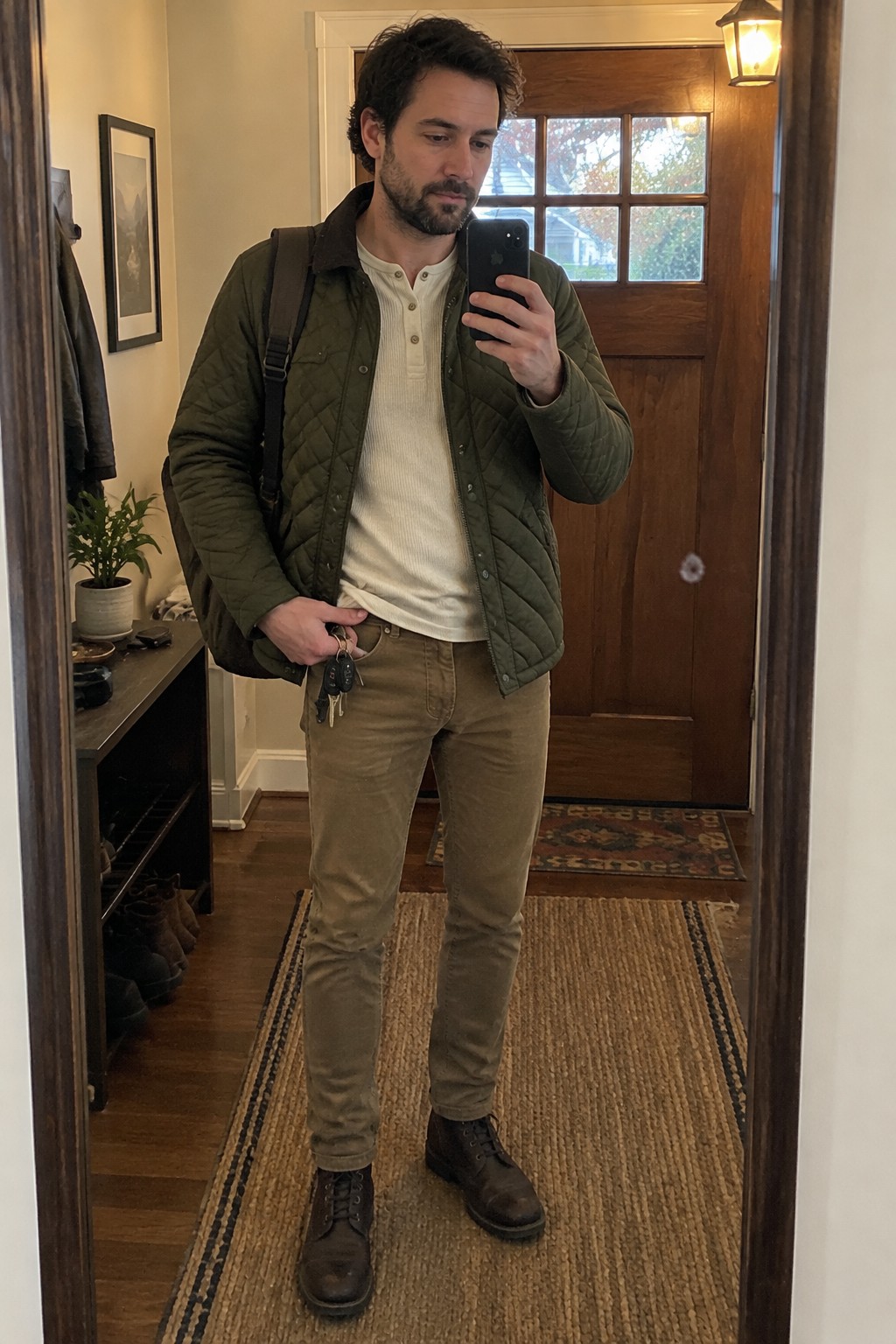 A man wearing an olive quilted jacket over a cream henley, tan-olive trousers, and dark brown lace-up boots, with a black shoulder bag.