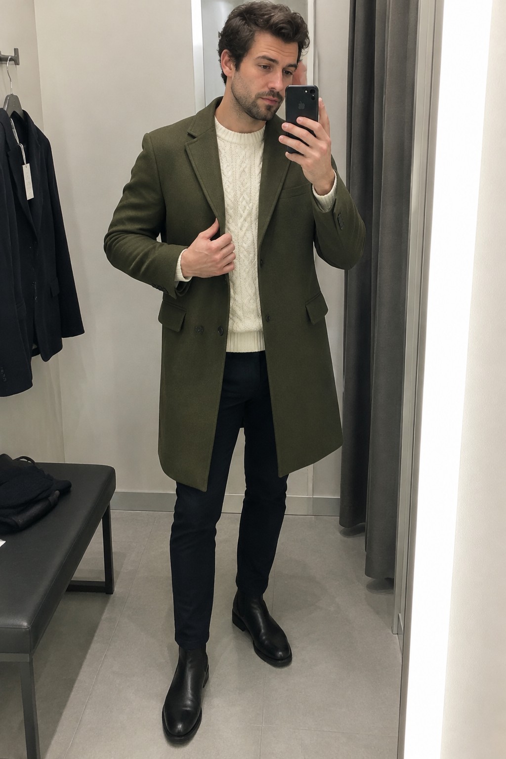 Man wearing a long olive wool coat over a cream cable-knit sweater with dark trousers and black Chelsea boots.