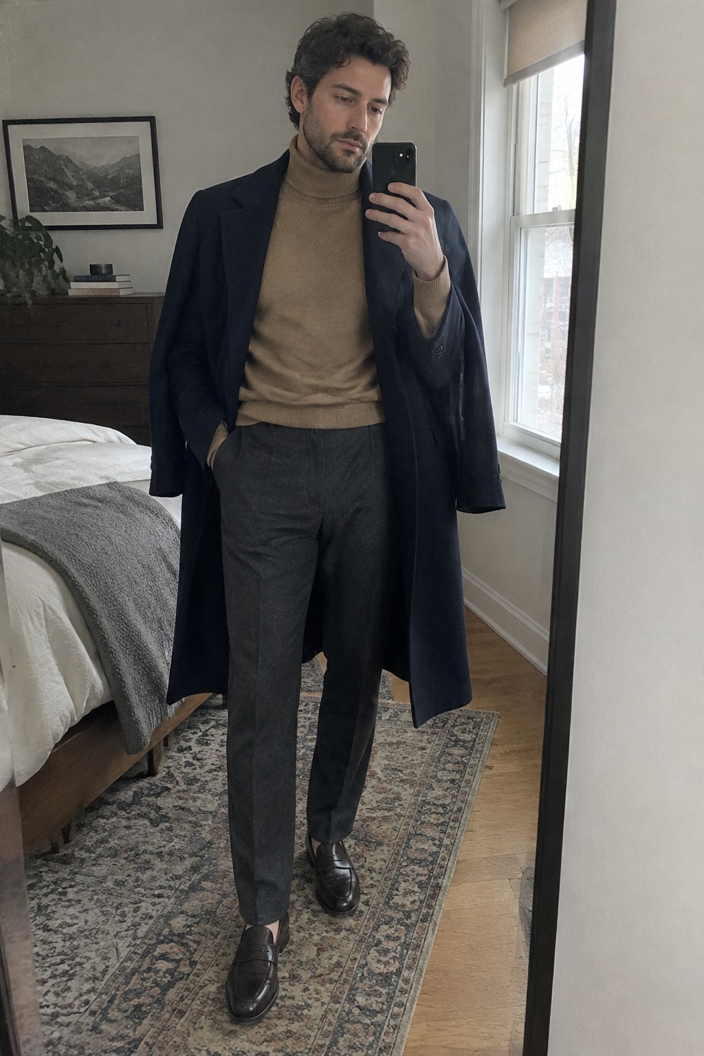 A man wearing a long navy overcoat over a camel turtleneck, charcoal tailored wool trousers, and dark brown loafers.