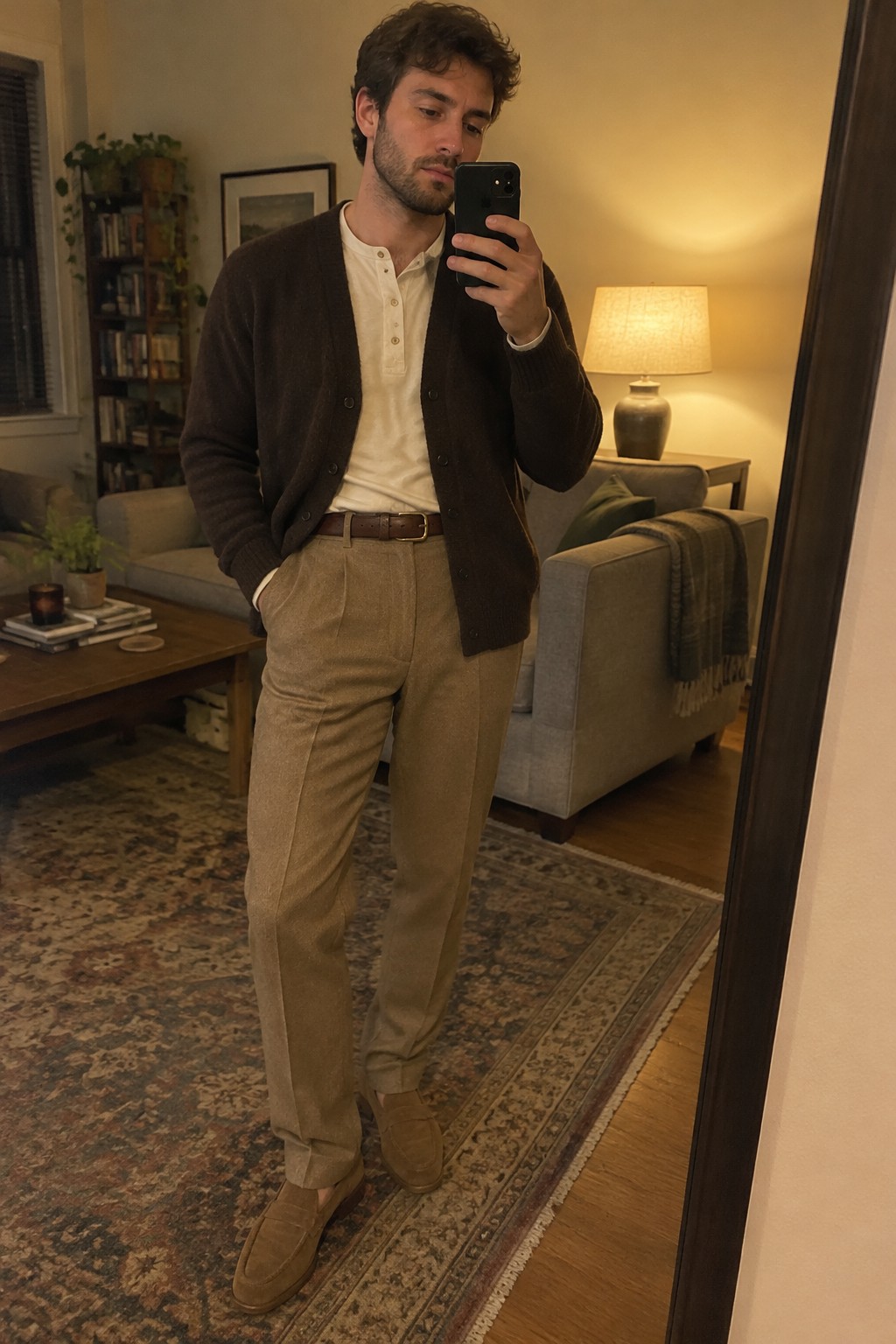 Man wearing a dark brown cardigan over a cream henley shirt, pleated taupe wool trousers, a brown leather belt, and brown suede loafers.