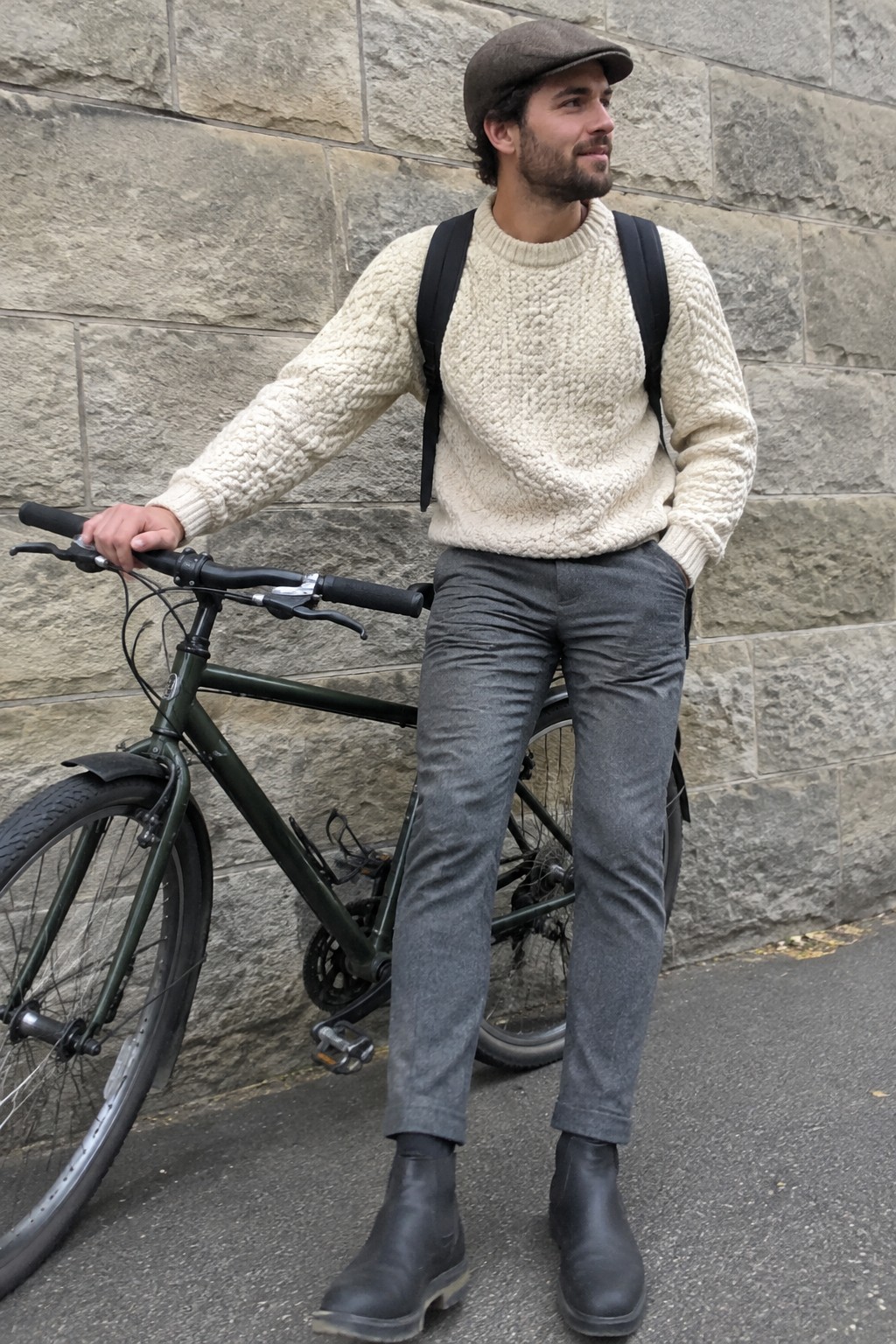 A man in a cream cable-knit sweater, grey tailored trousers, dark flat cap, black ankle boots, and black backpack stands beside a bicycle.