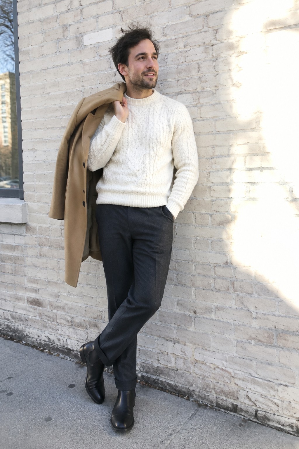 A man wearing a cream cable-knit sweater, charcoal wool trousers, black Chelsea boots, and a camel coat draped over his shoulder.