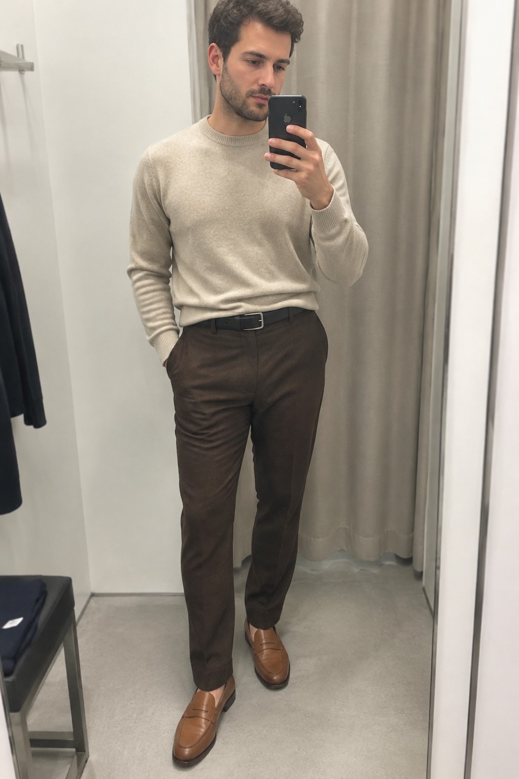 Man wearing a cream crewneck sweater tucked into dark brown tailored wool trousers with a black belt and brown leather loafers.