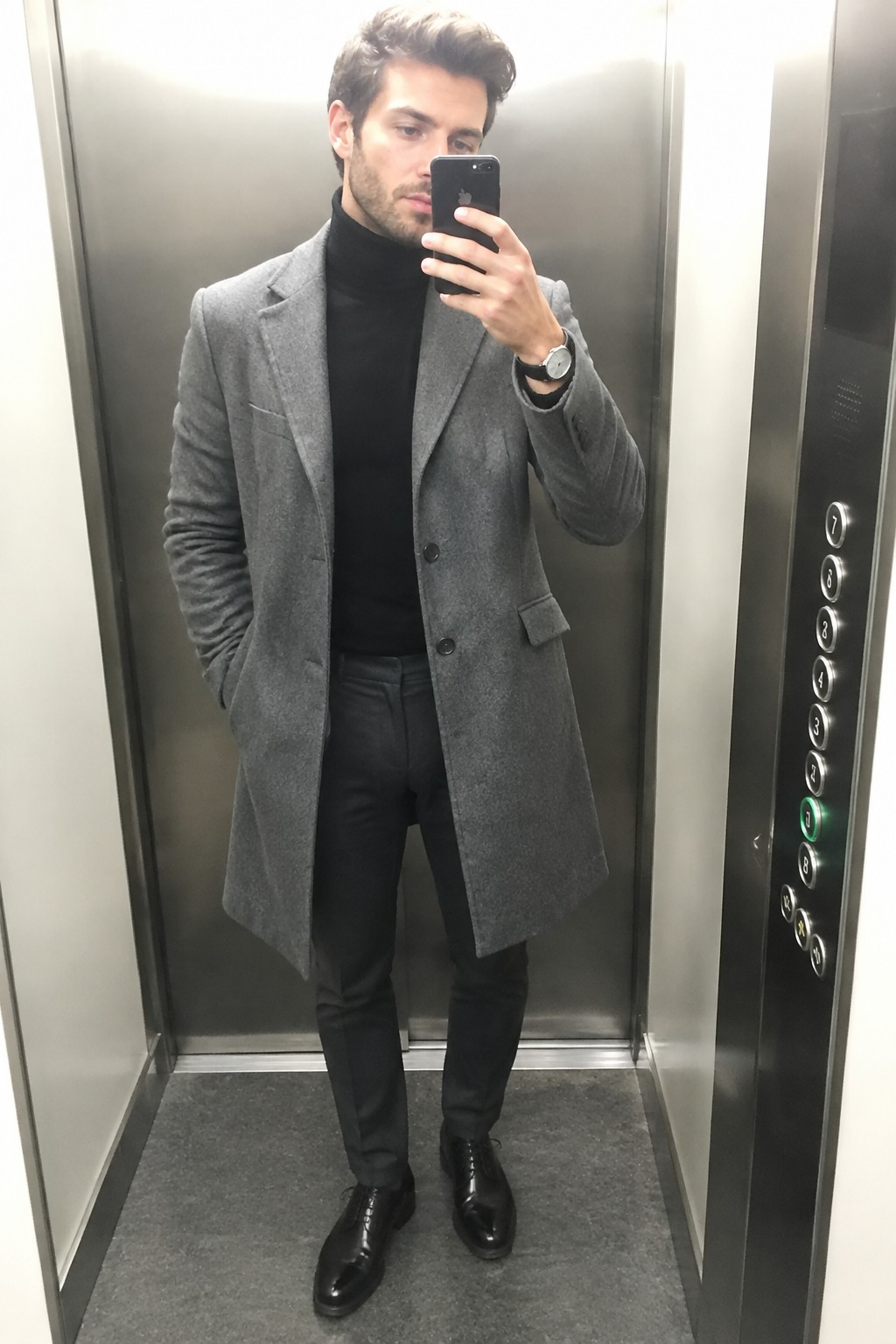 A man wearing a grey wool overcoat over a black turtleneck and black tailored trousers with black lace-up shoes and a wristwatch.