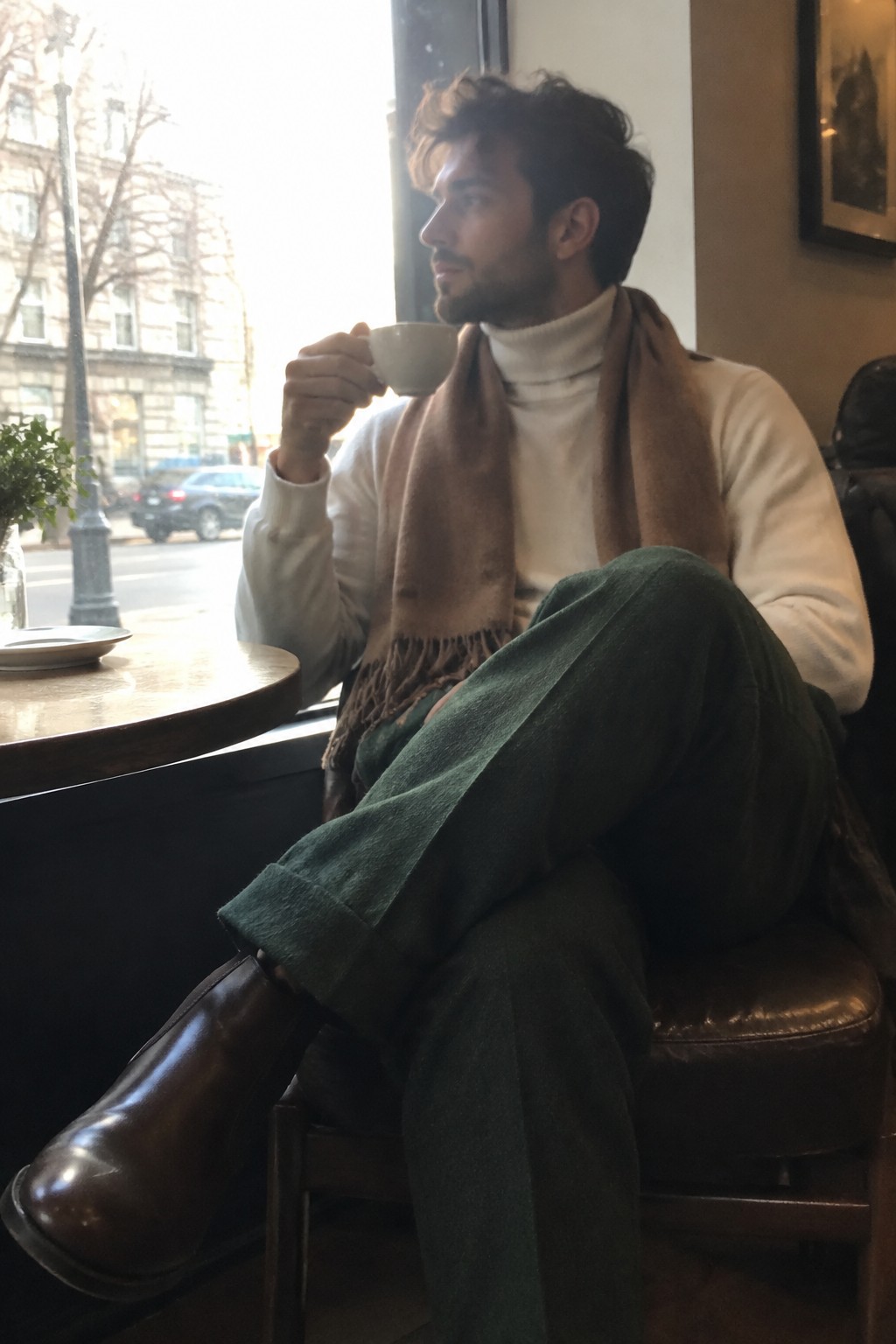 A man seated in a café wearing an ivory turtleneck, brown fringed scarf, deep green tailored wool trousers, and dark brown leather boots.