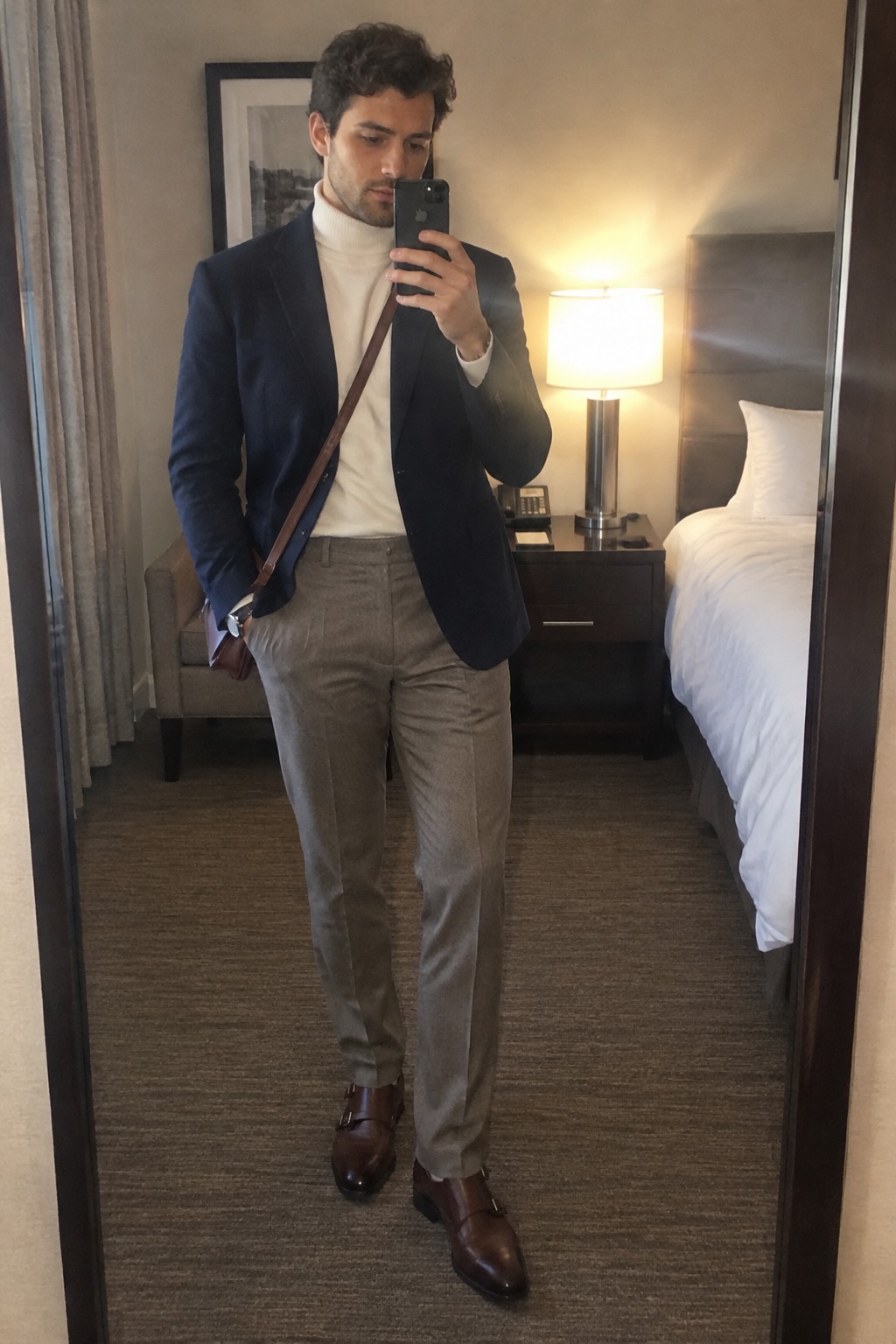 Man wearing a navy blazer over a cream roll-neck sweater with taupe wool trousers, brown double-monk shoes, and a brown crossbody bag.