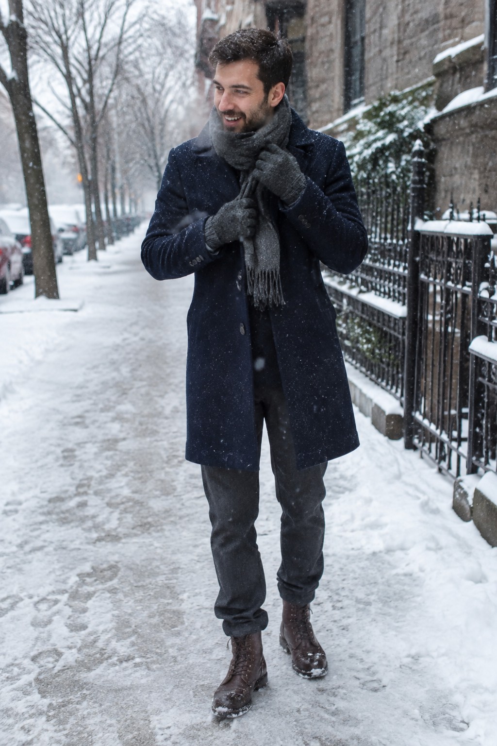 A man wears a long navy wool coat over dark trousers with a grey scarf and gloves and brown lace-up boots.
