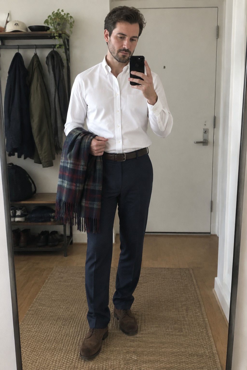 A man wears a white button-down shirt tucked into navy tailored wool trousers with a brown belt and brown suede shoes, holding a navy, green, and burgundy plaid scarf.