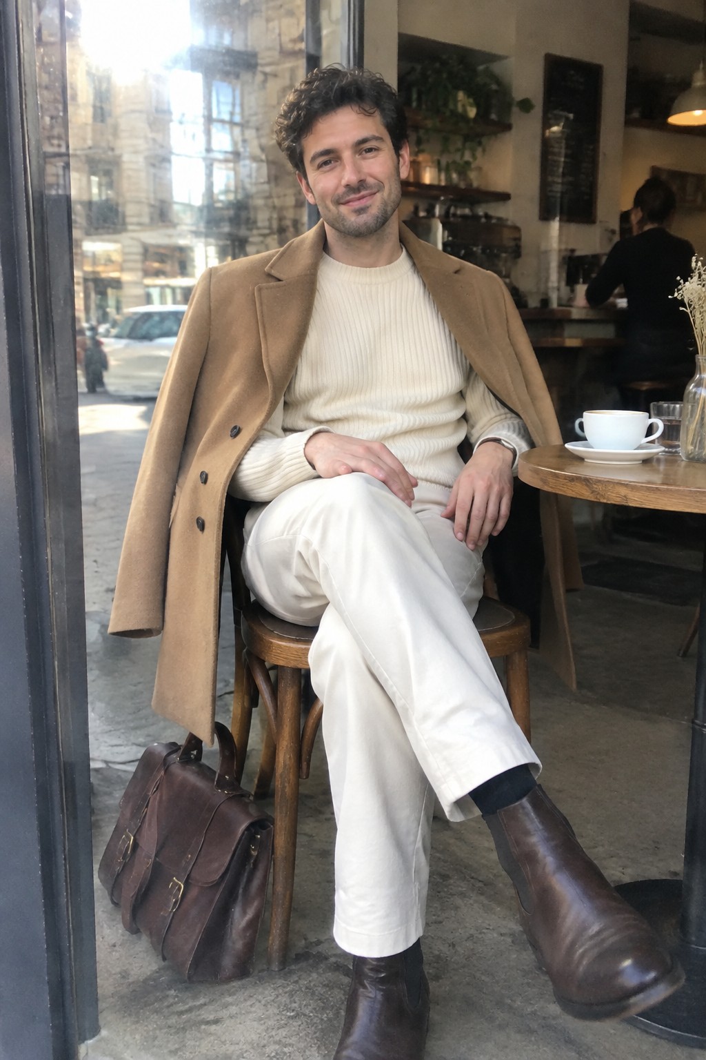 A man wears a camel overcoat over a cream ribbed sweater and cream trousers with dark brown leather ankle boots.