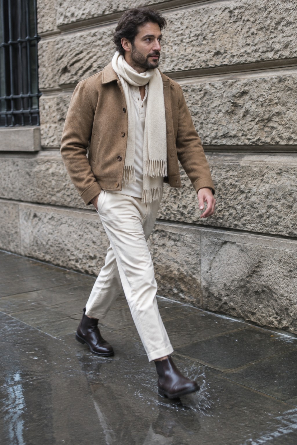 A man walks in a camel jacket, cream henley and scarf, cream trousers, and dark brown Chelsea boots.