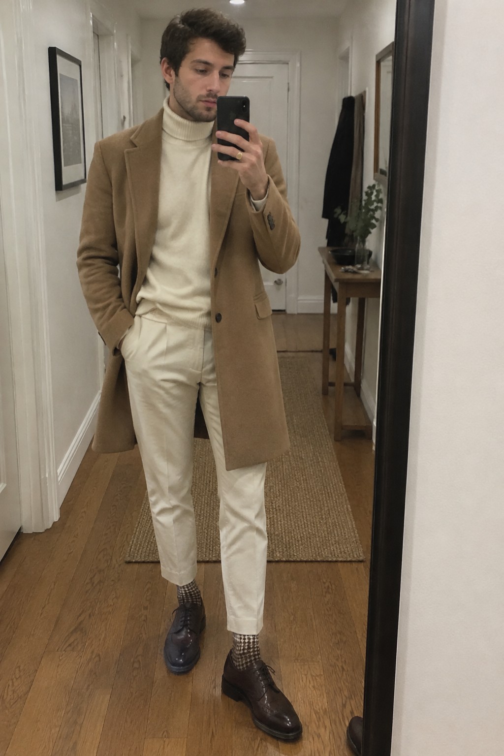 A man wears a camel overcoat over a cream ribbed turtleneck and cropped cream trousers, paired with brown leather oxford shoes and brown-and-cream checked socks.