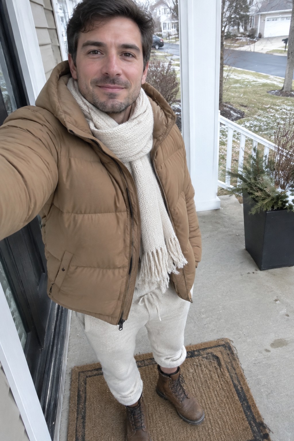 Man wearing a camel puffer jacket, cream scarf, cream knitwear and cuffed cream trousers with brown lace-up boots.