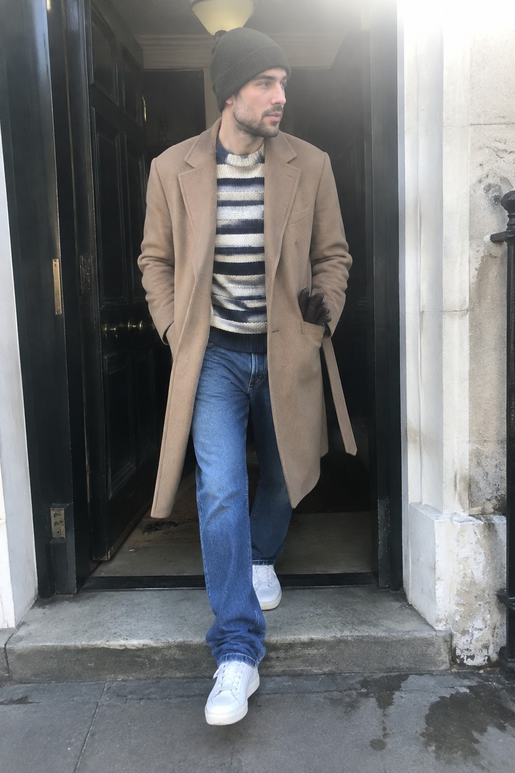Man wearing a long camel overcoat over a cream-and-navy striped sweater, blue jeans, a dark knit beanie, brown leather gloves, and white sneakers.