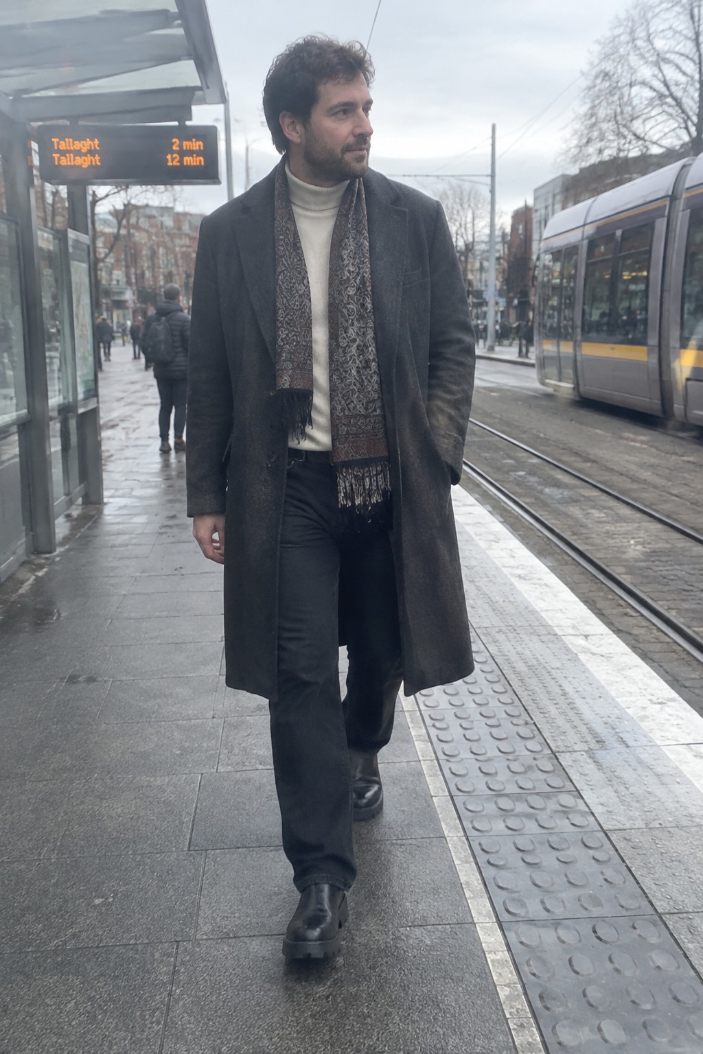 A man in a charcoal overcoat, cream turtleneck, patterned brown scarf, dark trousers, and black leather boots.