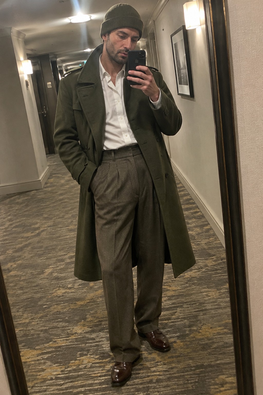 A man wears a long olive overcoat over a white shirt, pleated brown-gray trousers, a matching olive beanie, and dark brown loafers.