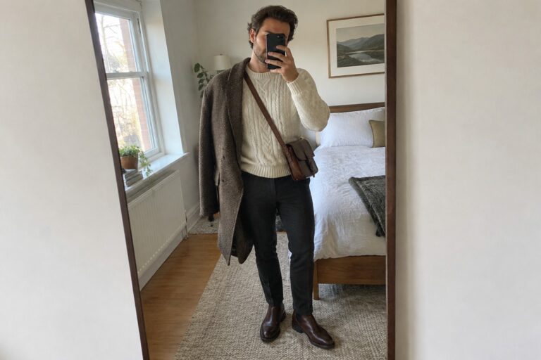 Man wearing a cream cable-knit sweater, charcoal trousers, brown Chelsea boots, and a gray-brown herringbone overcoat draped over one shoulder, with a brown leather crossbody bag.