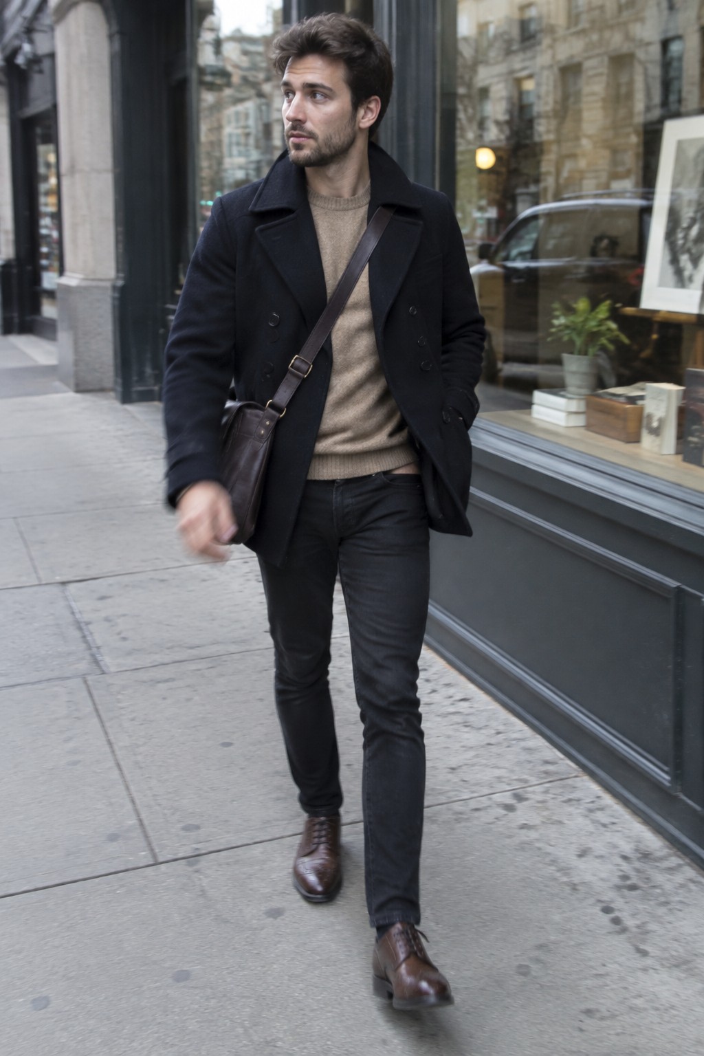A man wearing a black double-breasted peacoat over a taupe crewneck sweater, slim black jeans, brown leather lace-up boots, and a brown crossbody bag.