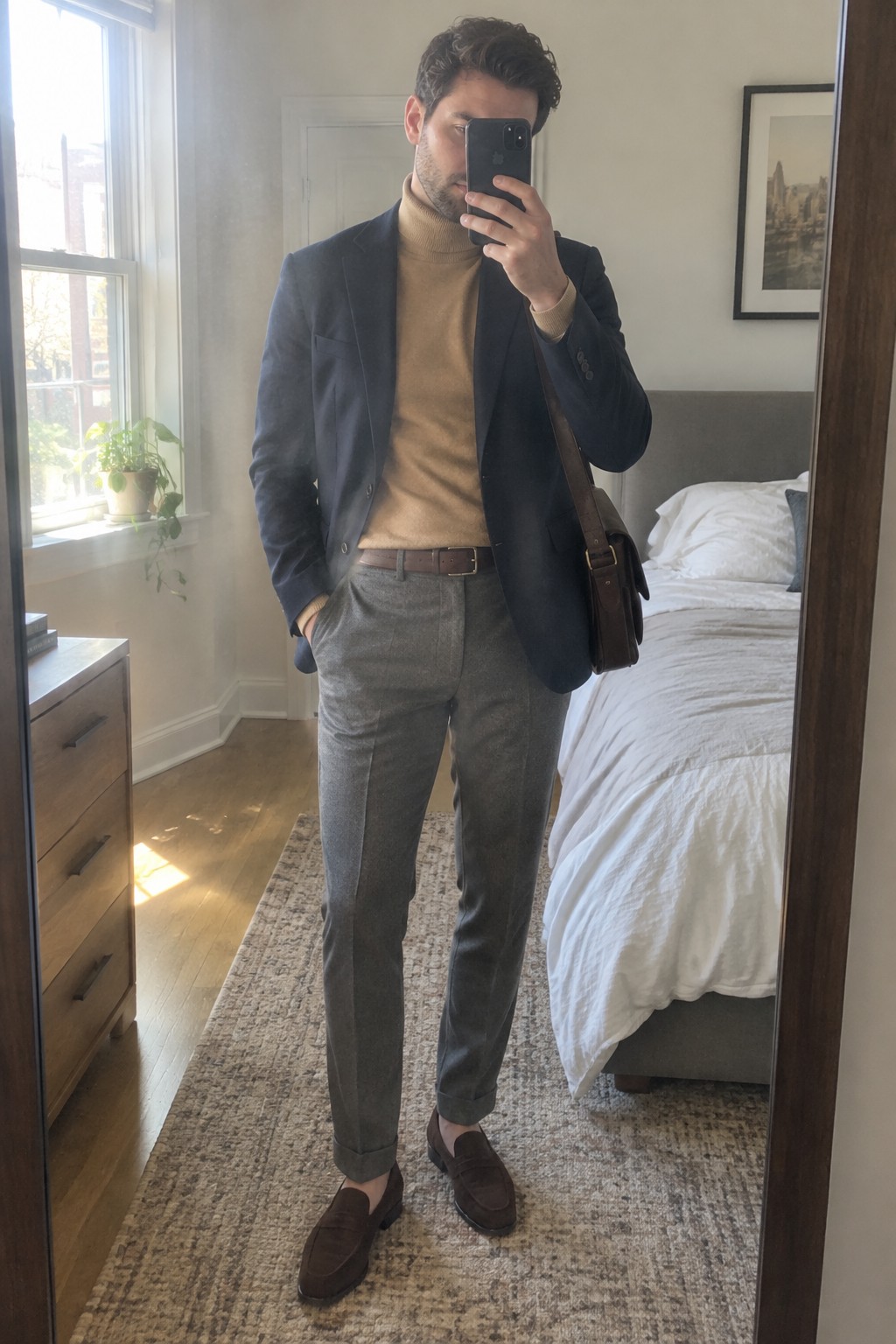 A man wearing a camel turtleneck, navy blazer, gray cuffed trousers, brown belt, brown suede loafers, and a brown leather shoulder bag.