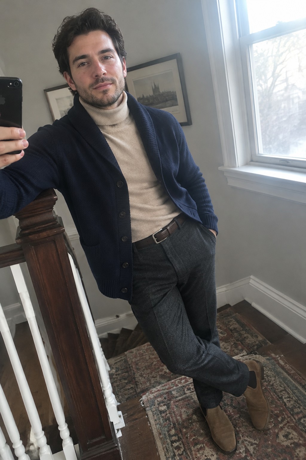 Man wearing a navy button-front cardigan over a cream turtleneck with charcoal trousers, a brown belt, and brown suede boots.