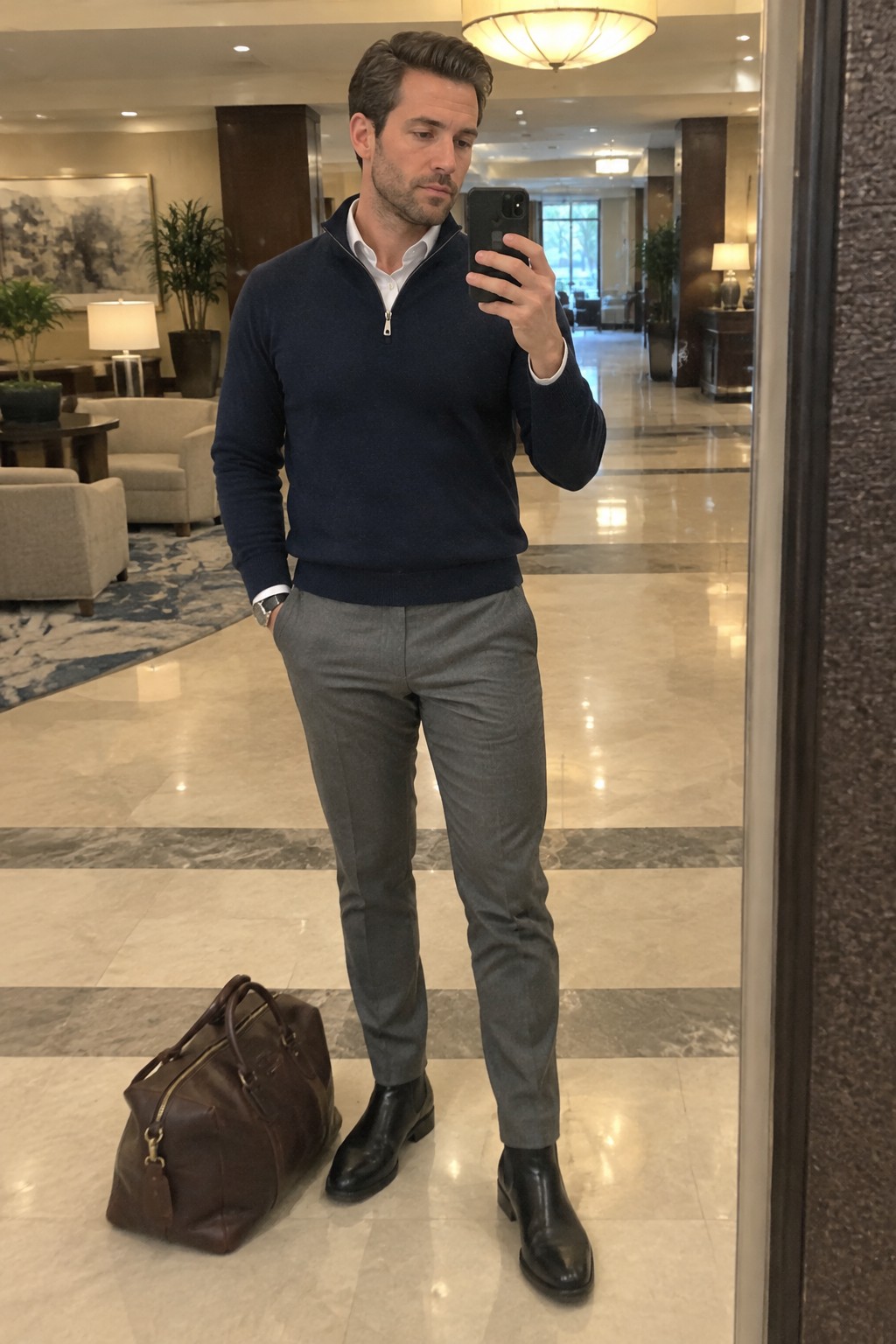 A man taking a mirror selfie in a navy quarter-zip sweater over a white shirt, gray trousers, black Chelsea boots, and a brown leather holdall.