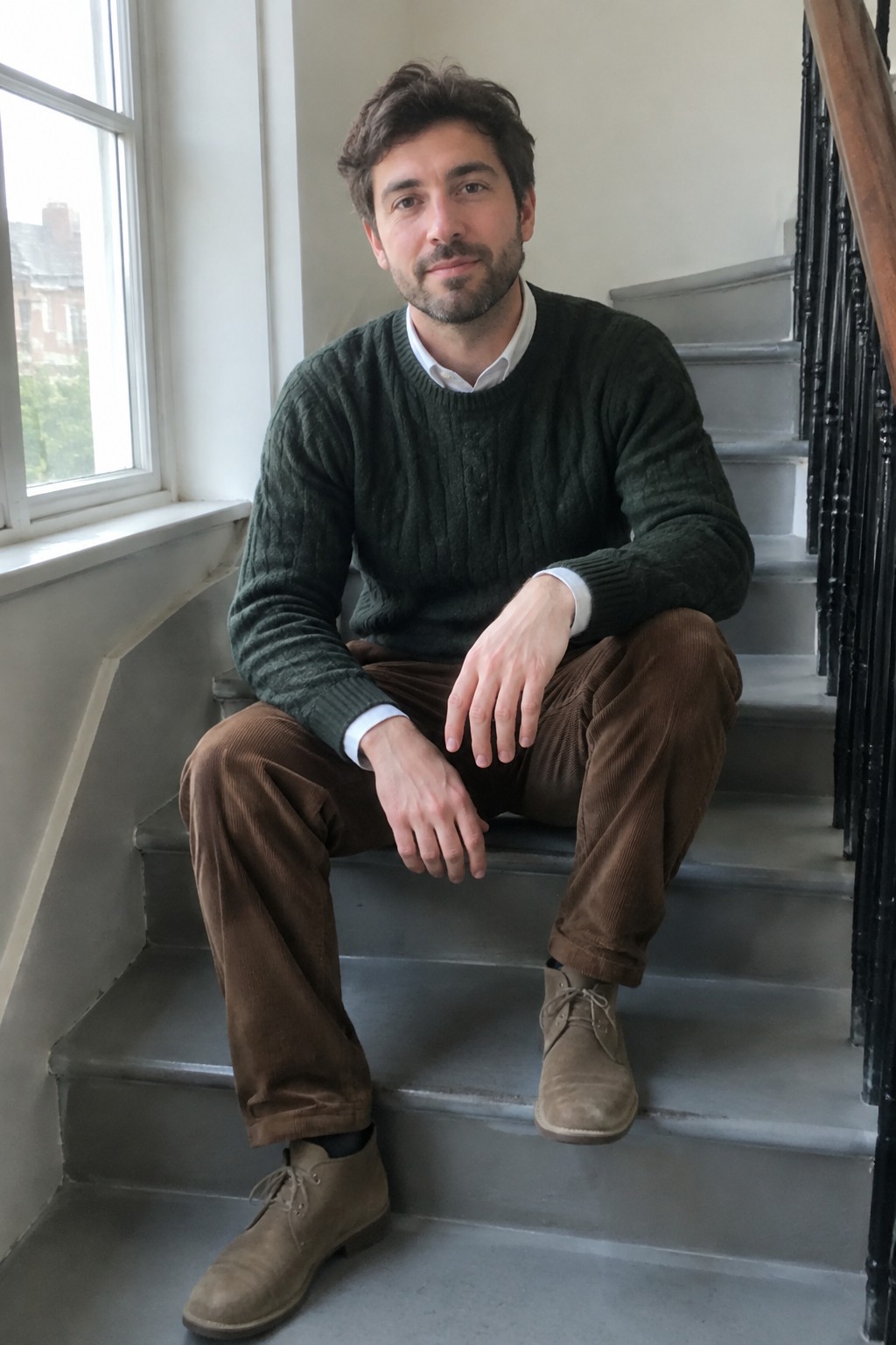 A man seated on stairs wearing a dark green cable-knit sweater over a white Oxford shirt, brown corduroy trousers, and tan suede lace-up boots.