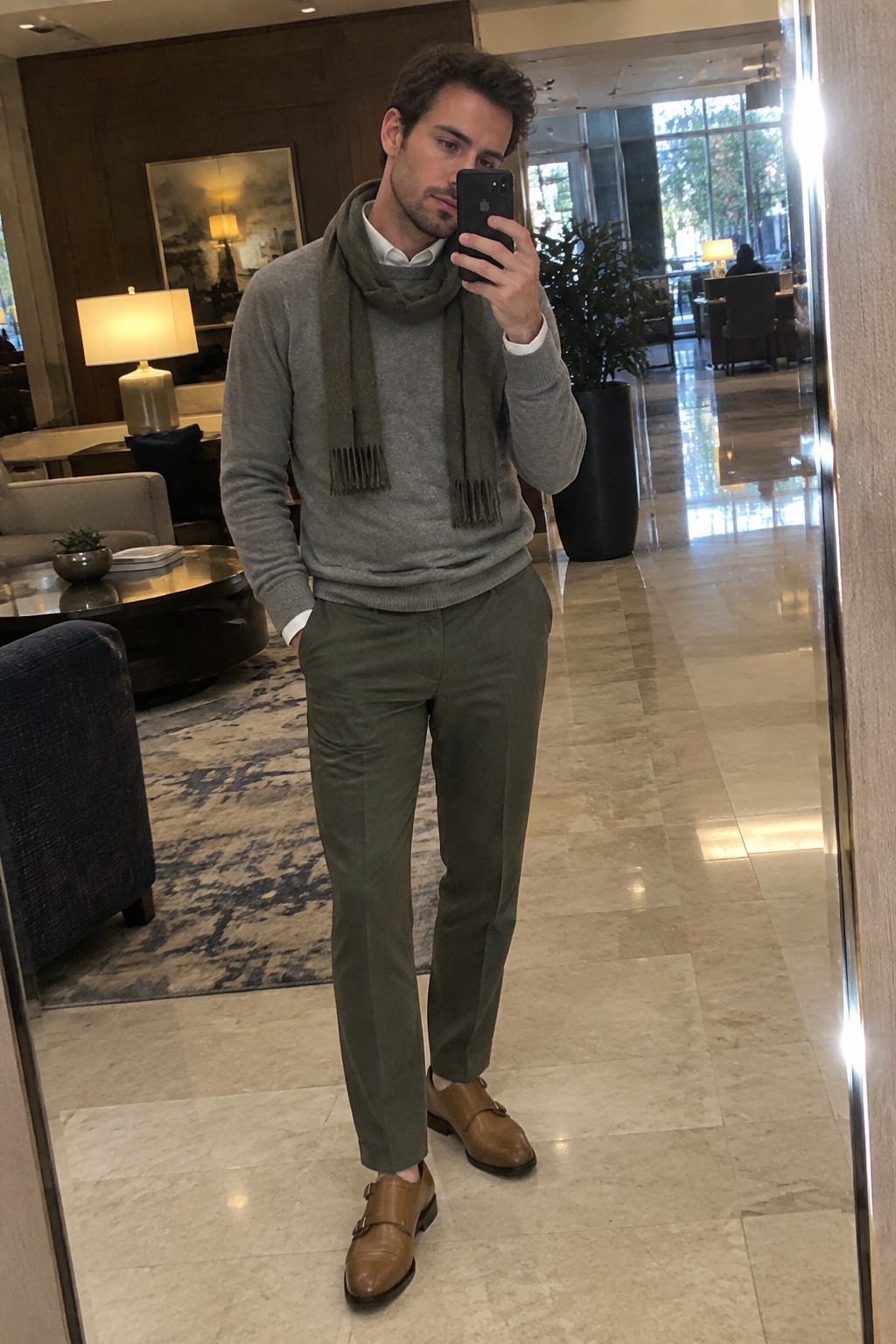 Man wearing a gray sweater over a white Oxford shirt, a gray scarf, olive trousers, and brown monk-strap shoes.