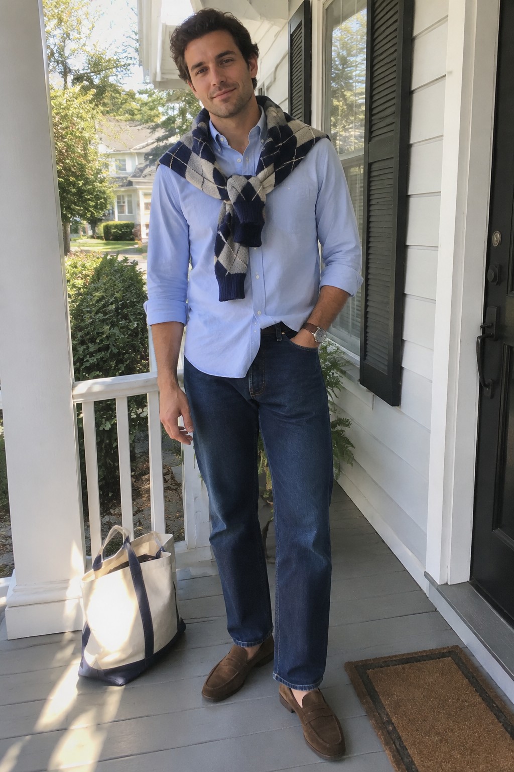 A man wearing a light blue Oxford shirt, an argyle sweater draped over his shoulders, dark jeans, brown suede loafers, and a watch.