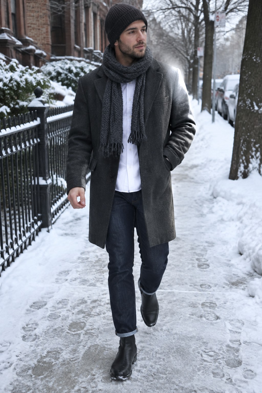 A man wears a white Oxford shirt under a charcoal overcoat and gray scarf, with dark cuffed jeans, black Chelsea boots, and a black knit beanie.