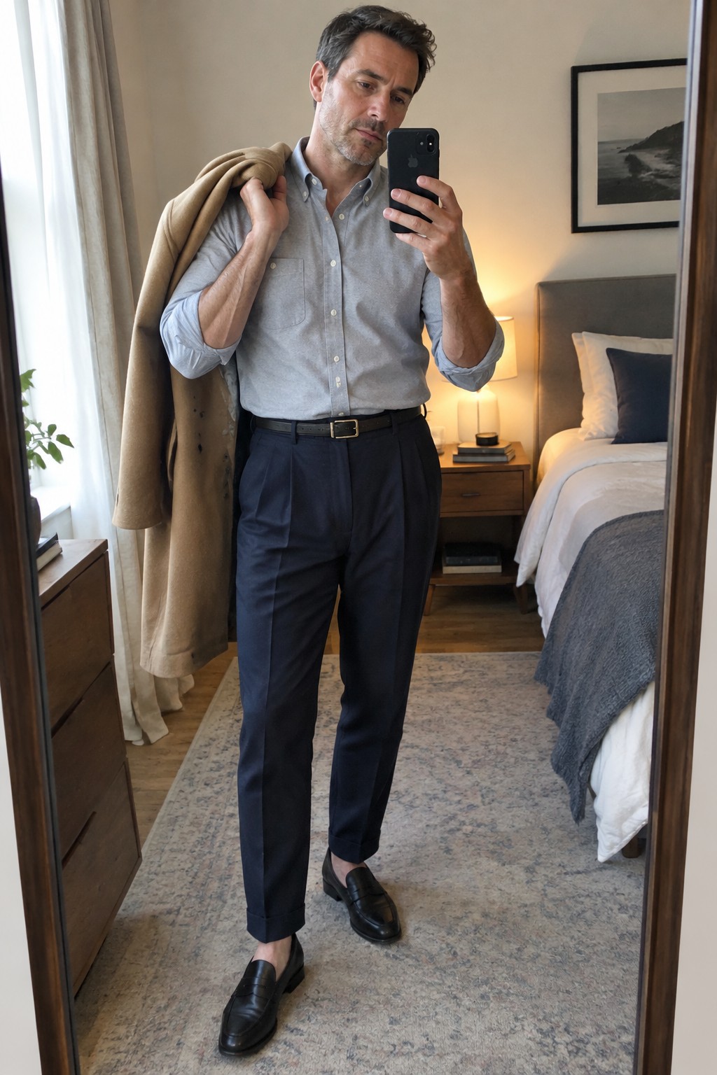 A man wears a pale blue-gray Oxford shirt tucked into navy pleated trousers with a black belt and black penny loafers, carrying a camel coat over one shoulder.