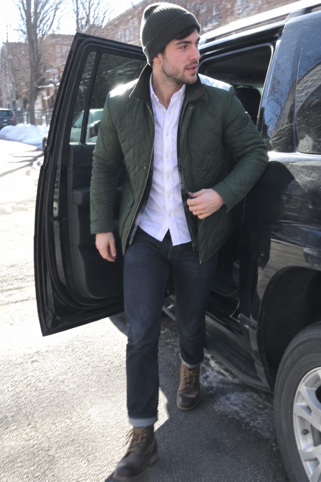 A man wearing a dark green quilted jacket over a white Oxford shirt, dark cuffed jeans, brown lace-up boots, and a dark knit beanie.