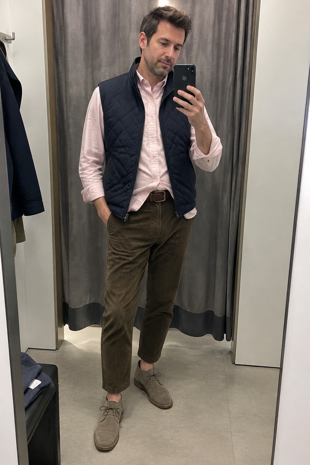 A man wears a pale pink Oxford shirt under an open navy quilted vest, with olive-brown corduroy trousers, a brown belt, and taupe suede lace-up shoes.