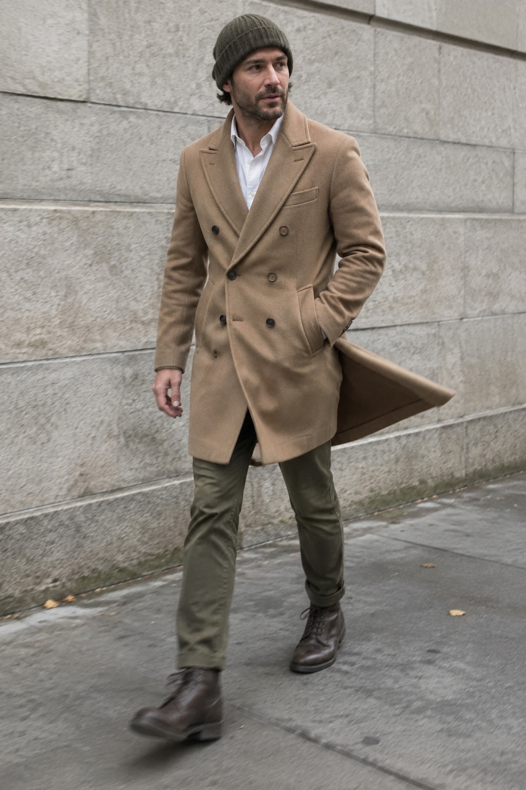 Man wearing a camel double-breasted overcoat over a white shirt, olive trousers, a dark ribbed beanie, and brown lace-up boots.