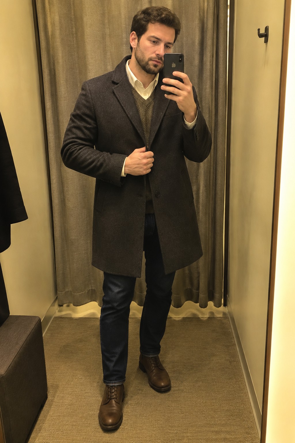 Man wearing a charcoal wool overcoat over a white shirt and brown V-neck sweater, with dark jeans and brown leather lace-up boots.