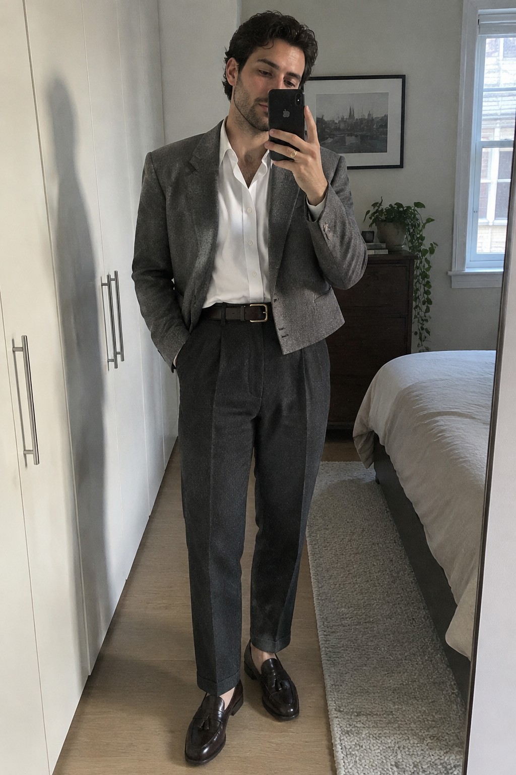 Man wearing a gray blazer, open-collar white shirt, charcoal pleated trousers, dark belt, and brown loafers.
