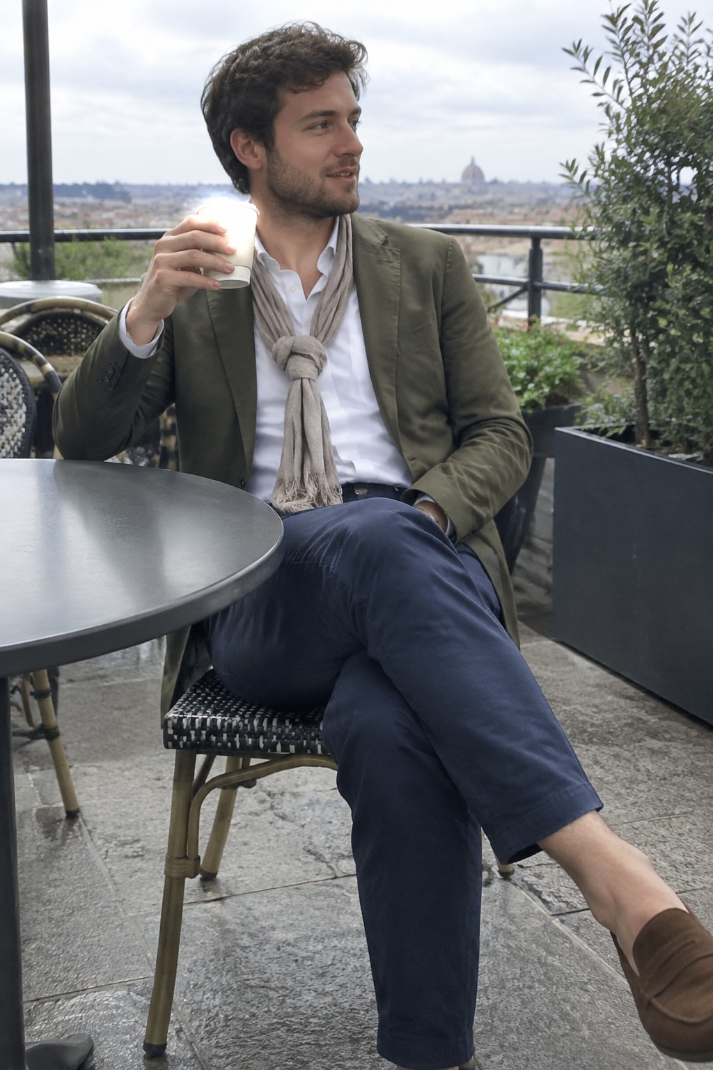 A man in an olive blazer, white shirt, taupe scarf, navy trousers, and brown suede loafers sits with a cup in hand.