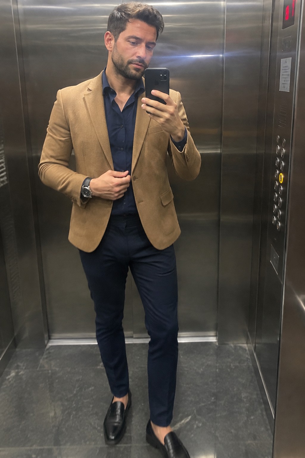 A man wears a camel blazer over a navy button-up shirt and navy trousers, with black loafers and a dark wristwatch.