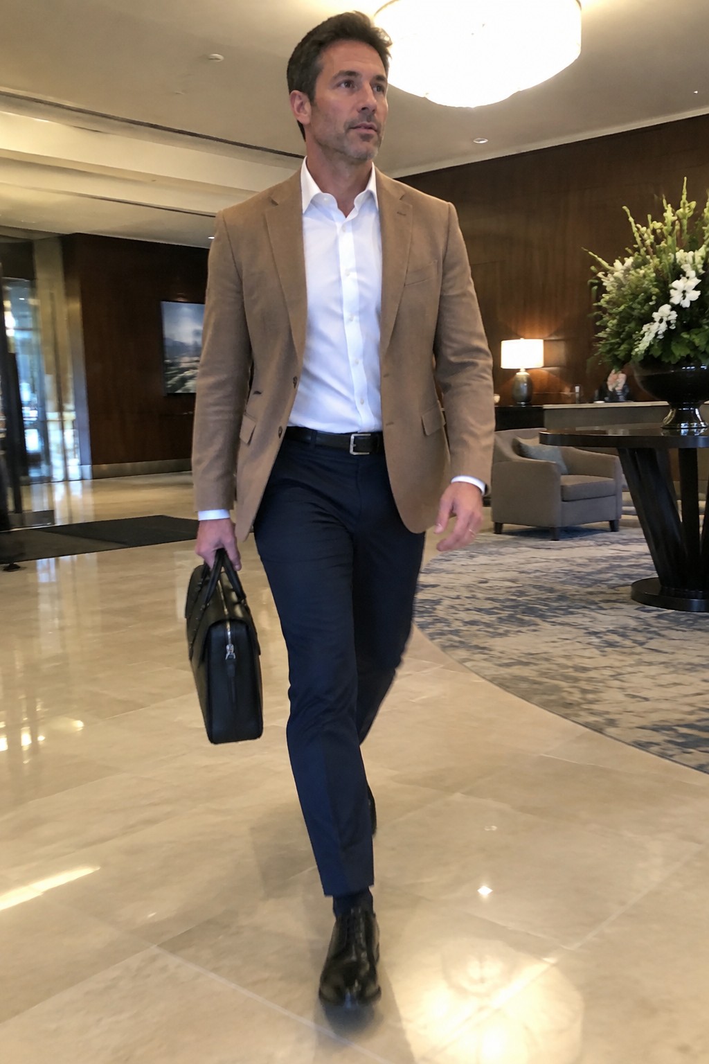 Man wearing a camel blazer, white open-collar shirt, navy trousers, black belt, black leather boots, and carrying a black briefcase.