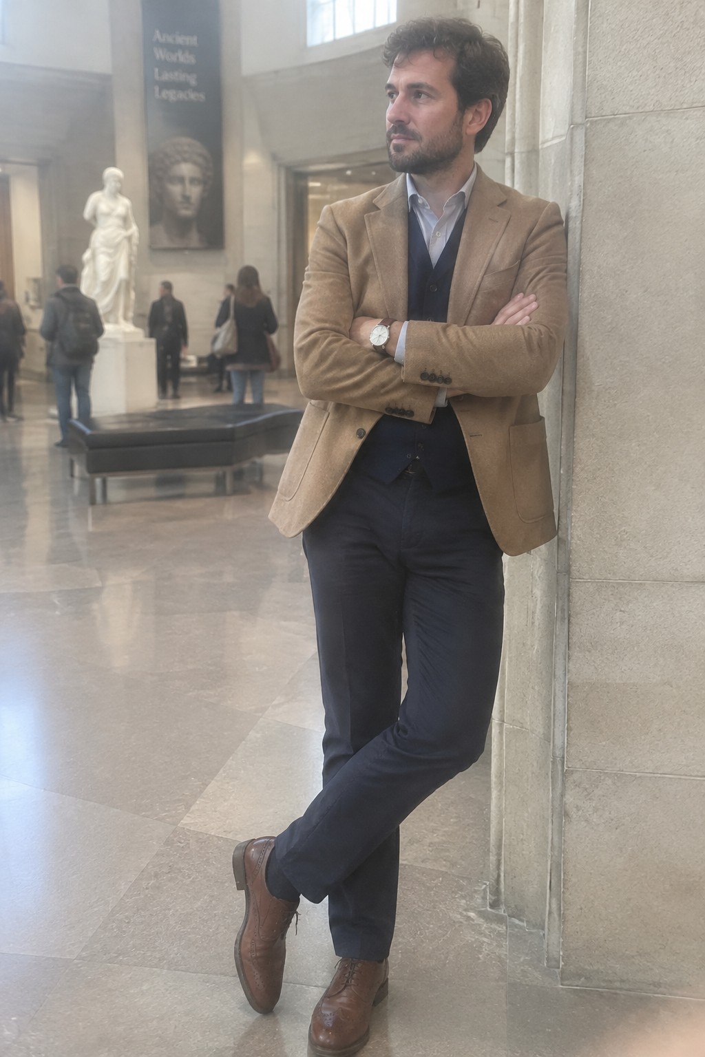 A man wearing a camel blazer, navy waistcoat and trousers, pale blue shirt, brown lace-up shoes, and a silver wristwatch.