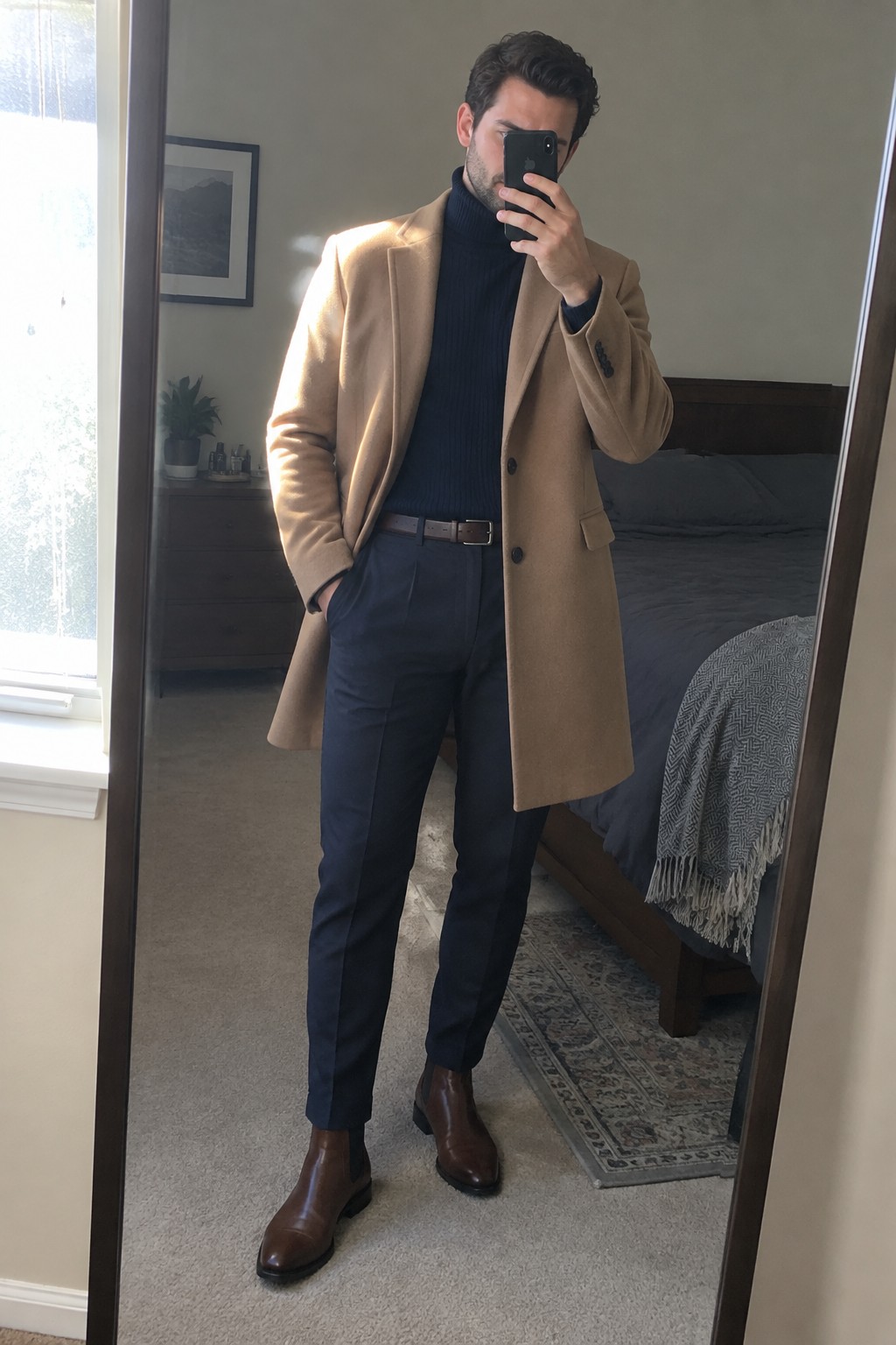 A man wears a camel overcoat over a navy ribbed turtleneck and navy trousers, finished with a brown leather belt and brown Chelsea boots.
